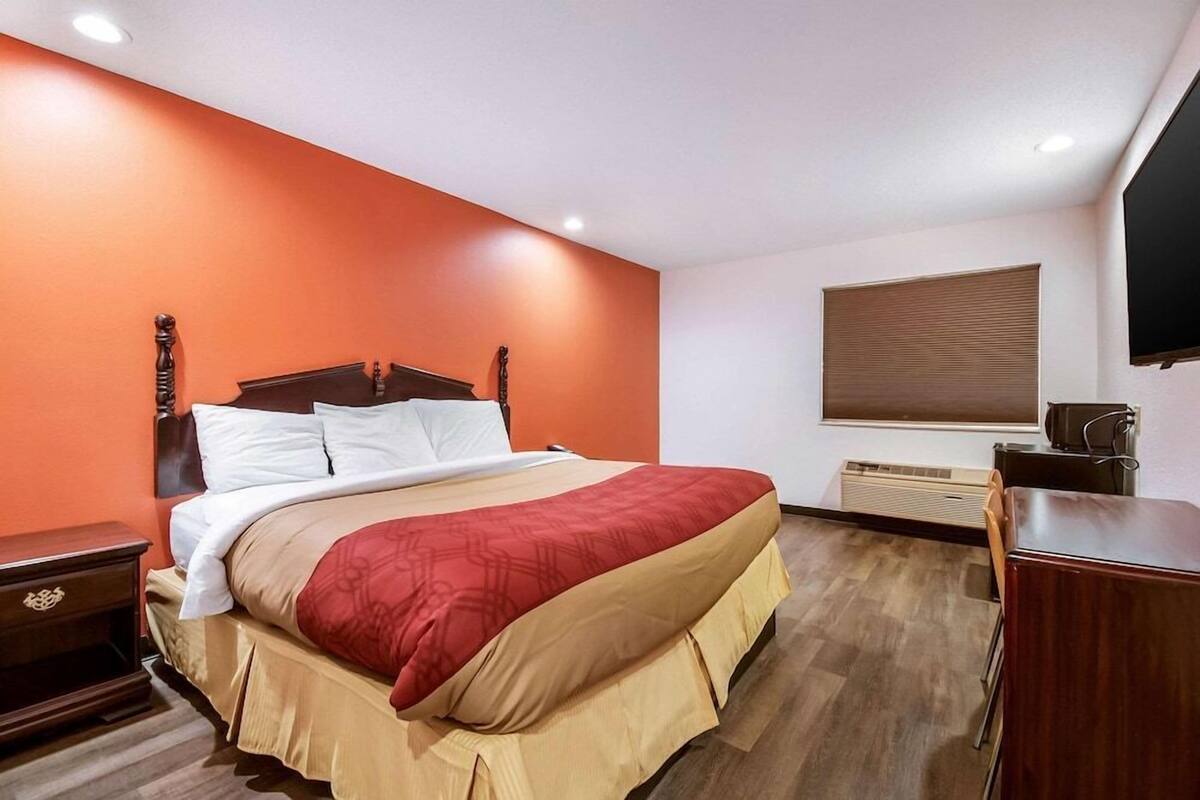 Habitación de hotel con cama, mesita de noche, escritorio, televisor y ventana con persianas.