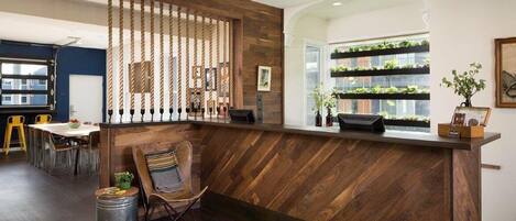 Zona de recepción con un mostrador de madera, un área de asientos con sillas, una mesa de comedor y vista a una pared verde con plantas.