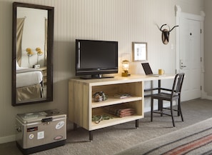 A hotel room with a wooden desk, a television, a lamp, a chair, a suitcase, and a mirror.