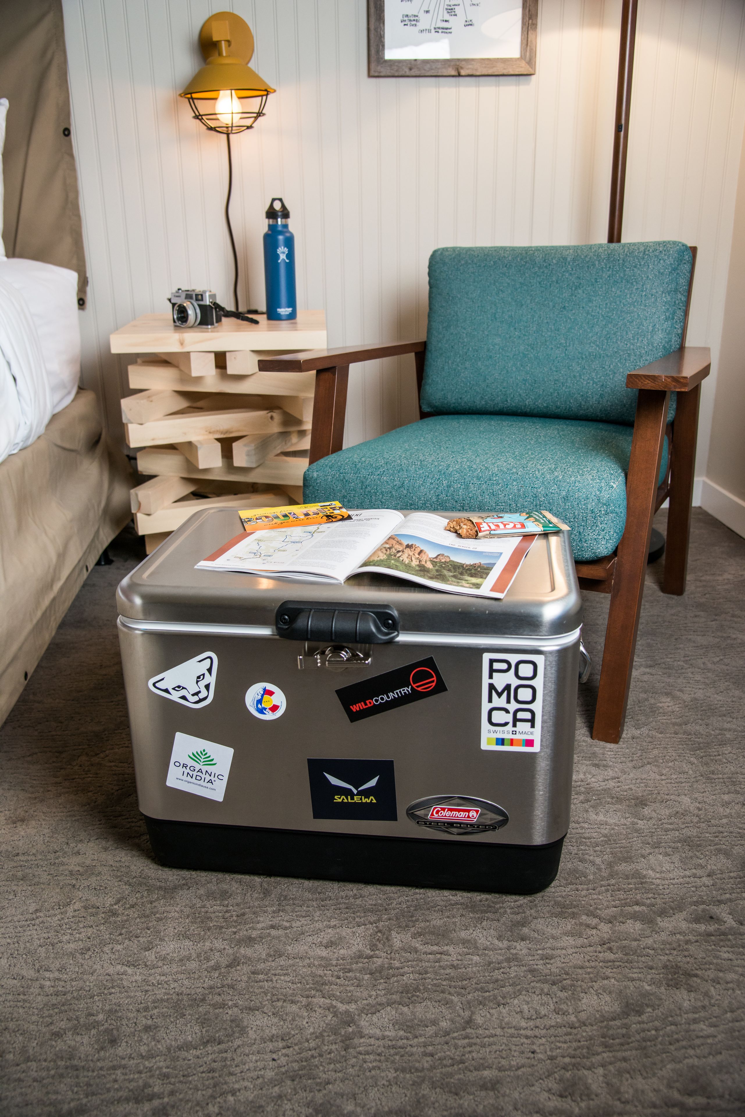 A suitcase with travel stickers, a teal chair, a wooden side table, a lamp, and a bed in a hotel room.