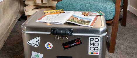 A suitcase with travel stickers, a teal chair, a wooden side table, a lamp, and a bed in a hotel room.