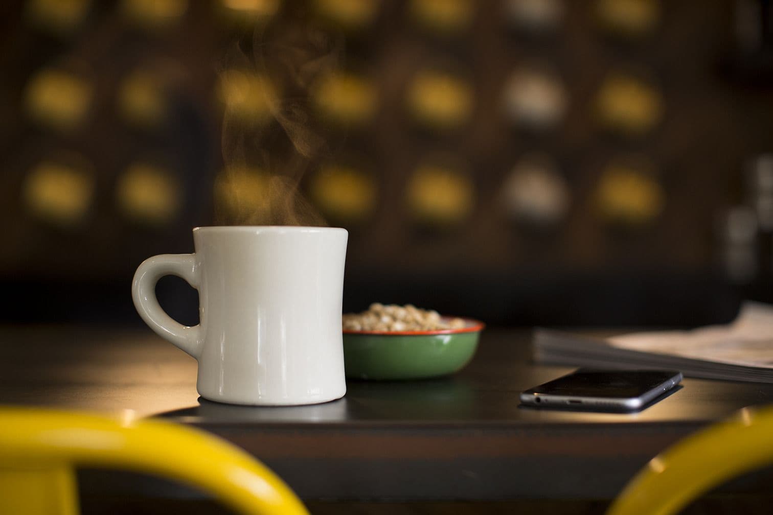 Eine weiße Tasse mit Dampf, eine grüne Schüssel mit Müsli und ein Smartphone auf einem Tisch.