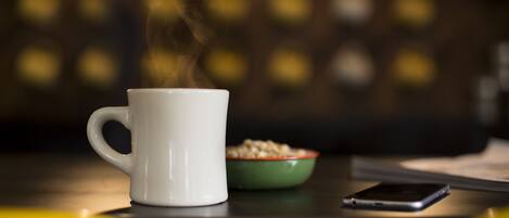 A white mug with steam, a green bowl of cereal, and a smartphone on a table.