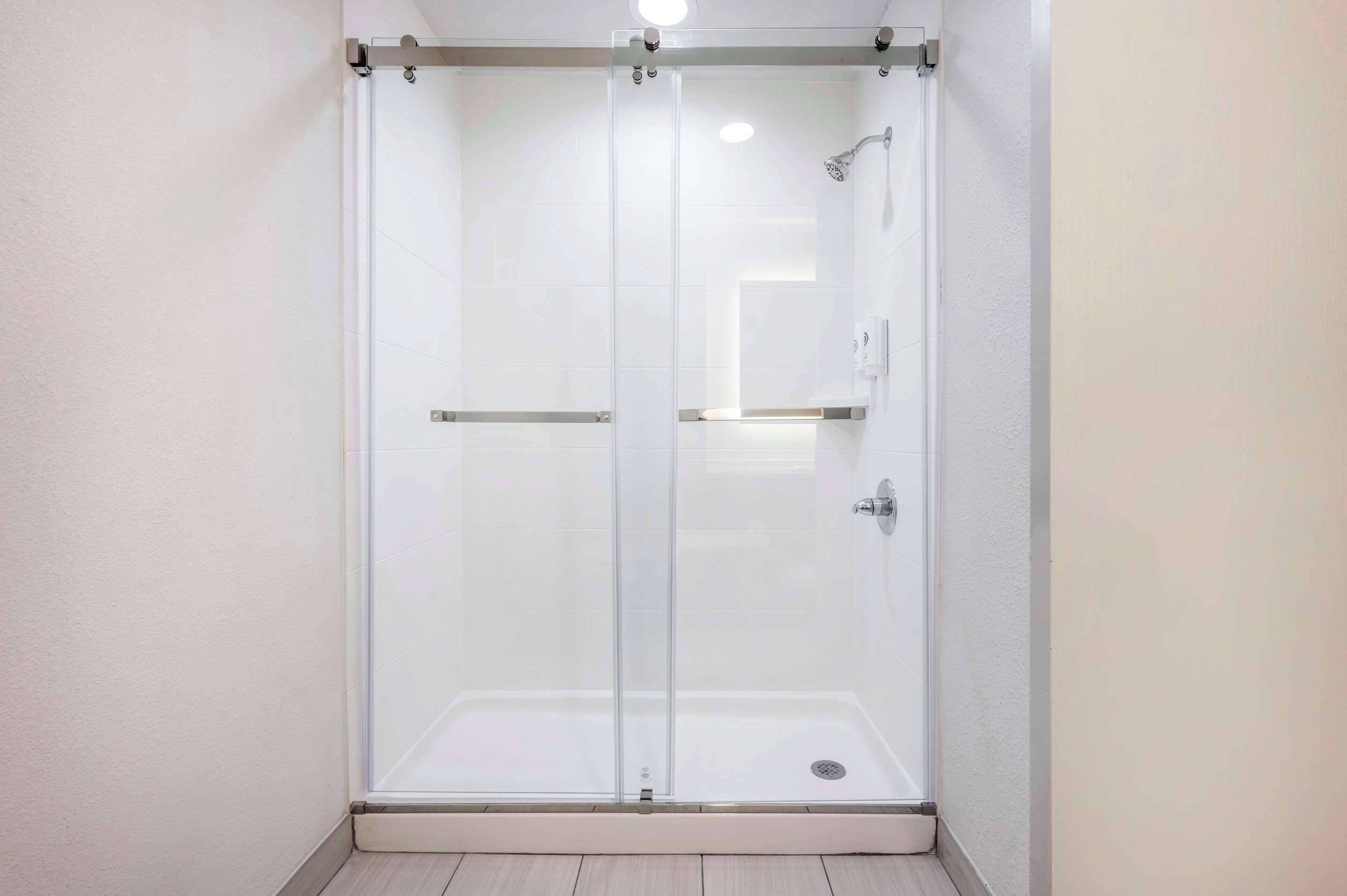 A glass-enclosed shower area with a white tiled floor and walls, a handheld showerhead, and a control panel.