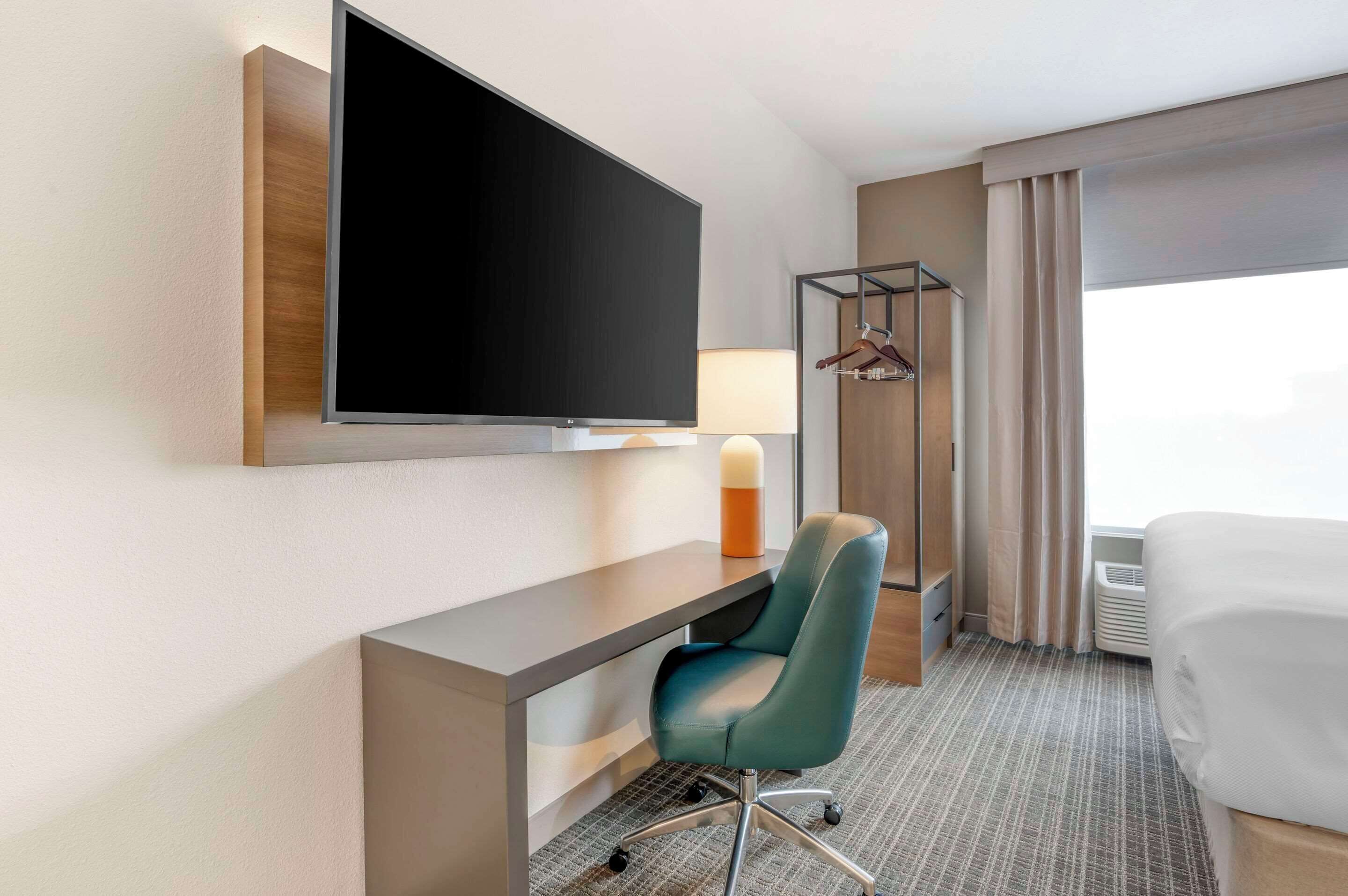 Habitación de hotel con un televisor de pantalla plana grande montado en la pared, un escritorio con una silla, una mesita de noche con una lámpara, una cama y una ventana con cortinas.