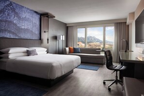 A modern hotel room with a large bed, a desk, a chair, and a view of the mountains.