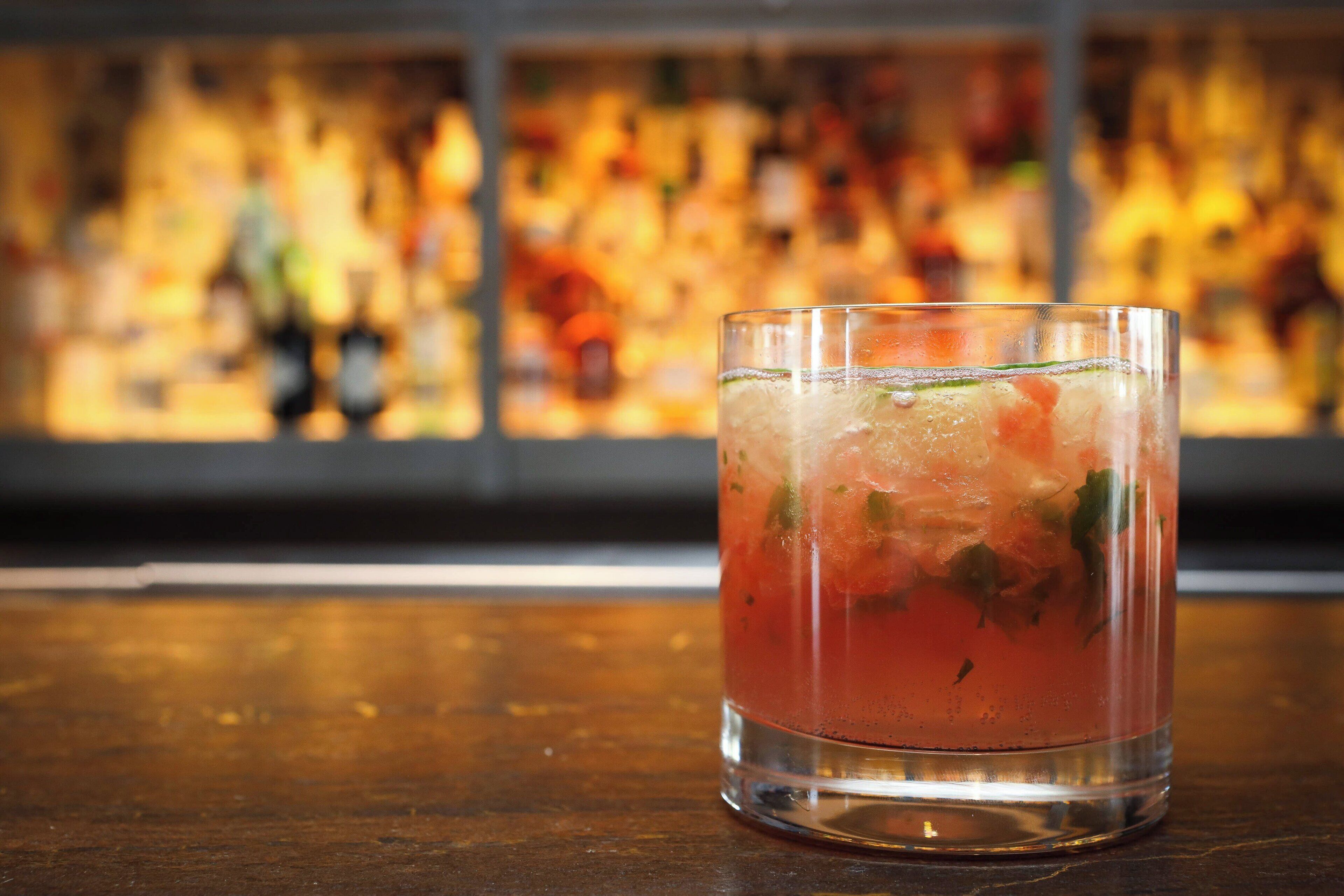 A glass of iced beverage on a wooden surface with a blurred background of bottles.