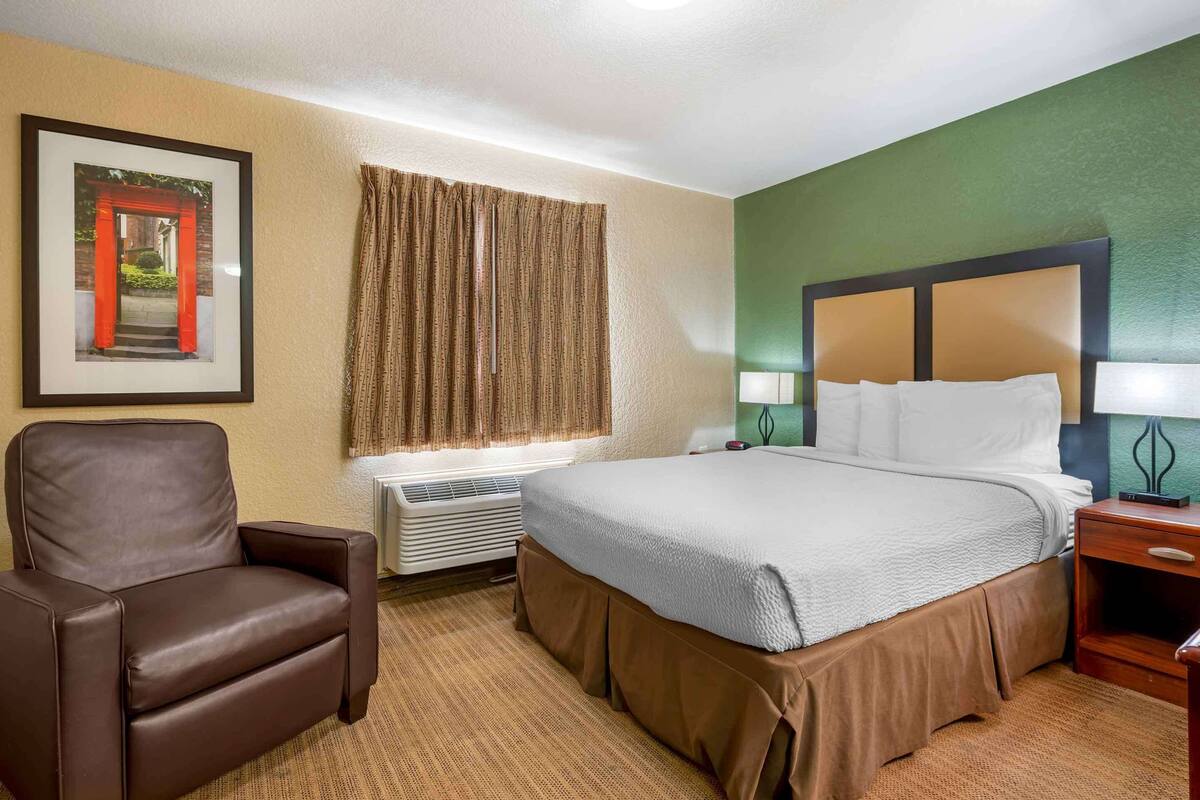A hotel room with a bed, a chair, a nightstand, a lamp, a framed picture on the wall, and a window with curtains.