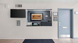 A reception desk with a 'WELCOME' sign, a flat-screen TV, a digital kiosk, and a door with a sign indicating office hours. - Extended Stay America Suites Fort Wayne North (Fort Wayne)