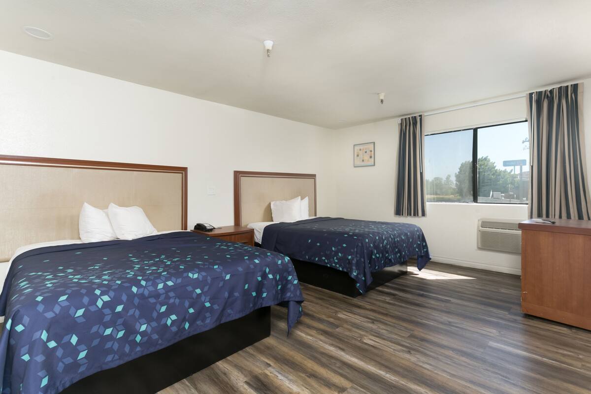 A hotel room with two beds, a wooden floor, a desk, and a window with curtains.