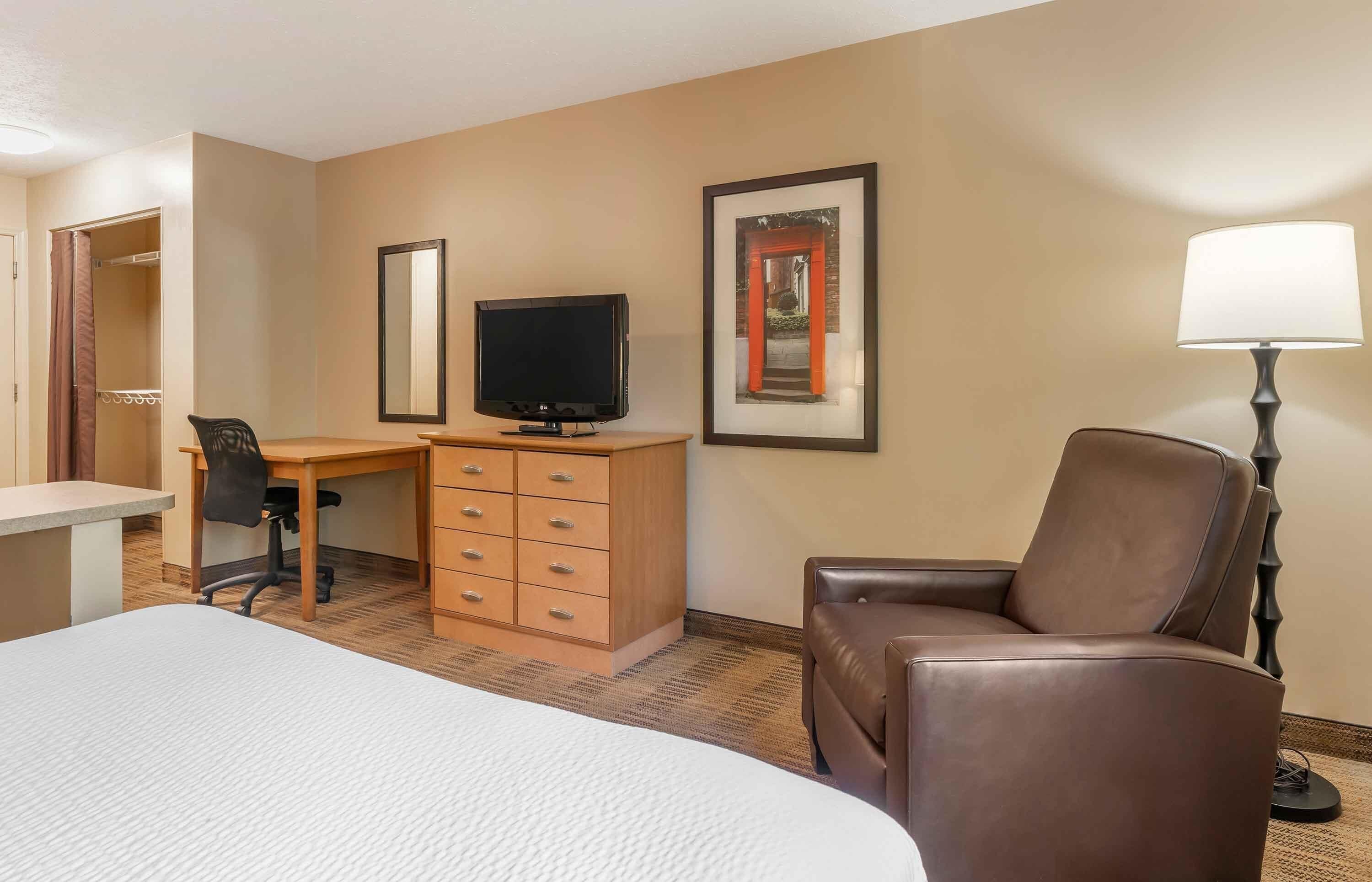 A hotel room with a bed, a desk with a television, a chair, a dresser, and a lamp.