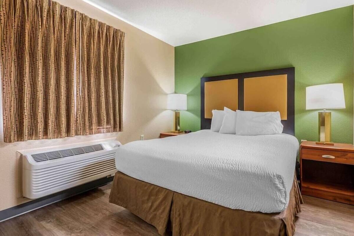 Habitación de hotel con una cama grande, dos mesitas de noche con lámparas, aire acondicionado en la pared y una cortina en la ventana.
