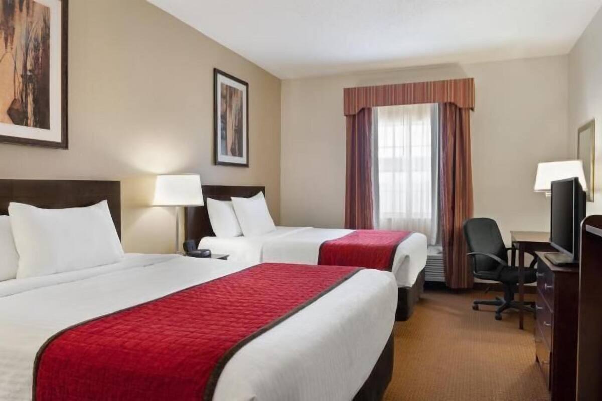 Quarto de hotel com duas camas, uma escrivaninha com cadeira, televisão e uma janela com cortinas.