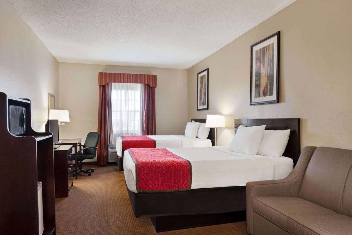 Quarto de hotel com duas camas, uma escrivaninha, uma cadeira, uma televisão e um sofá.
