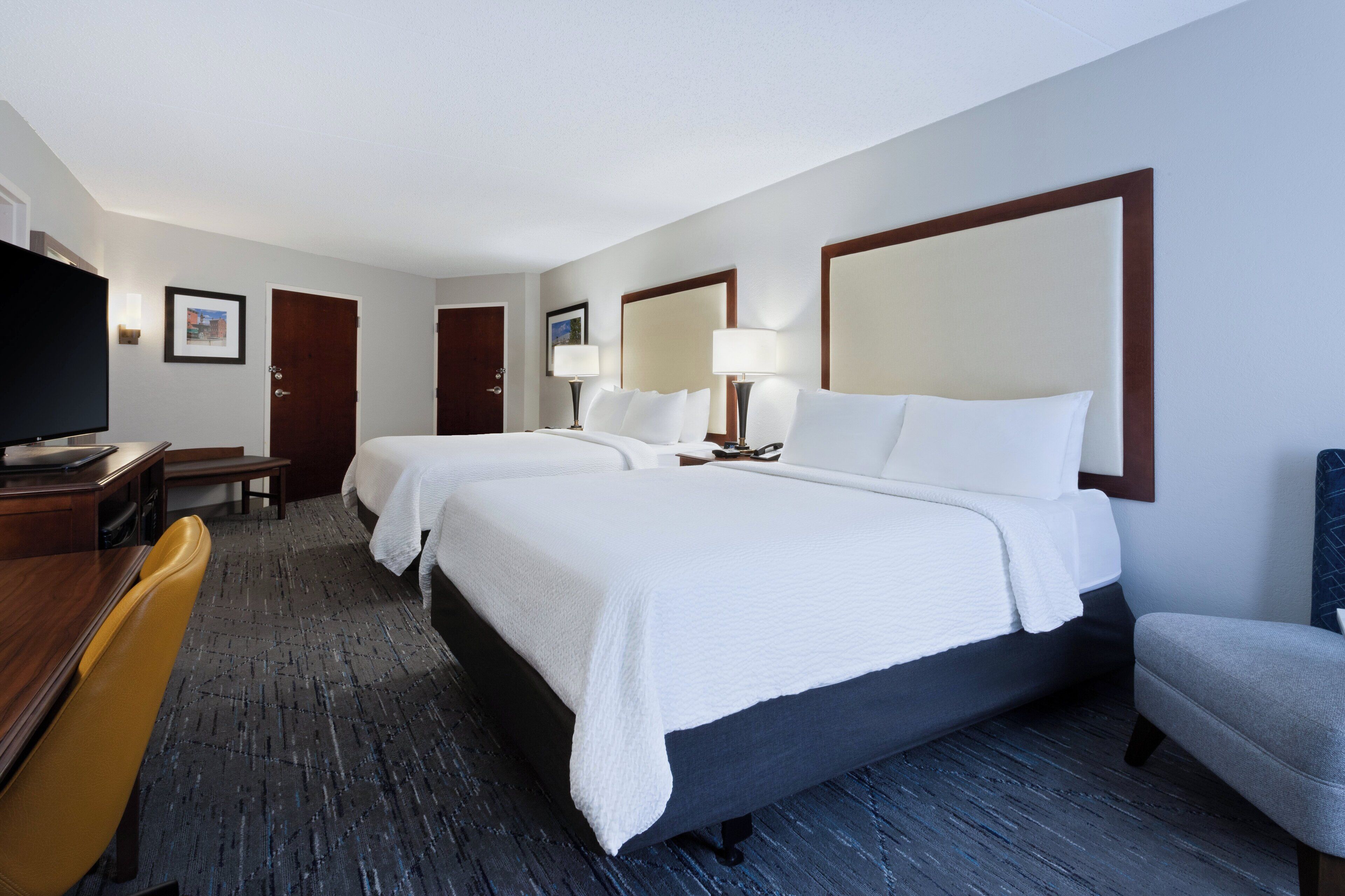 Habitación de hotel con dos camas, un escritorio con silla, un televisor y un espejo grande.