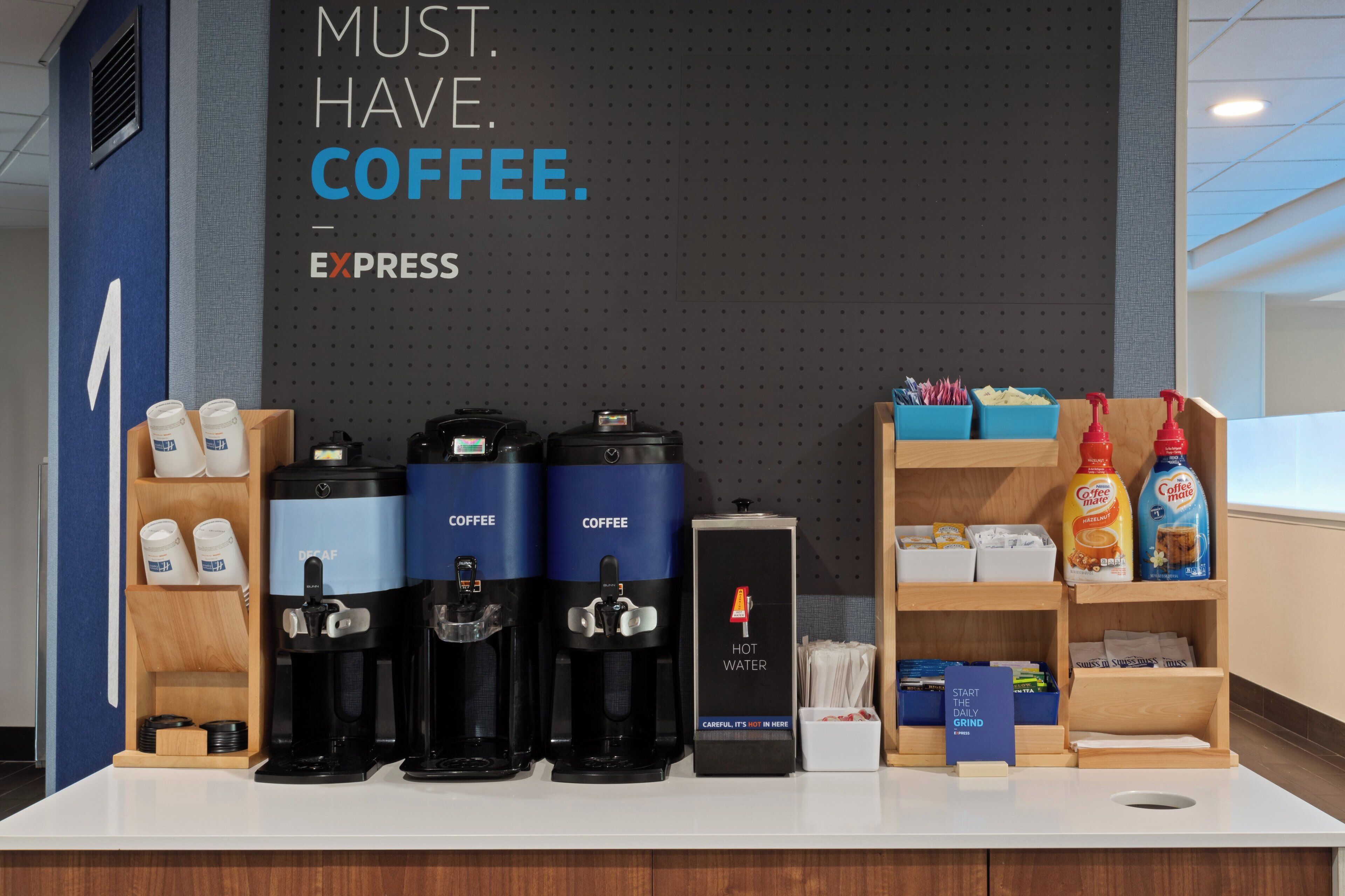 Estação de café com várias máquinas de café e um aviso que diz 'PRECISO. DE. CAFÉ'.