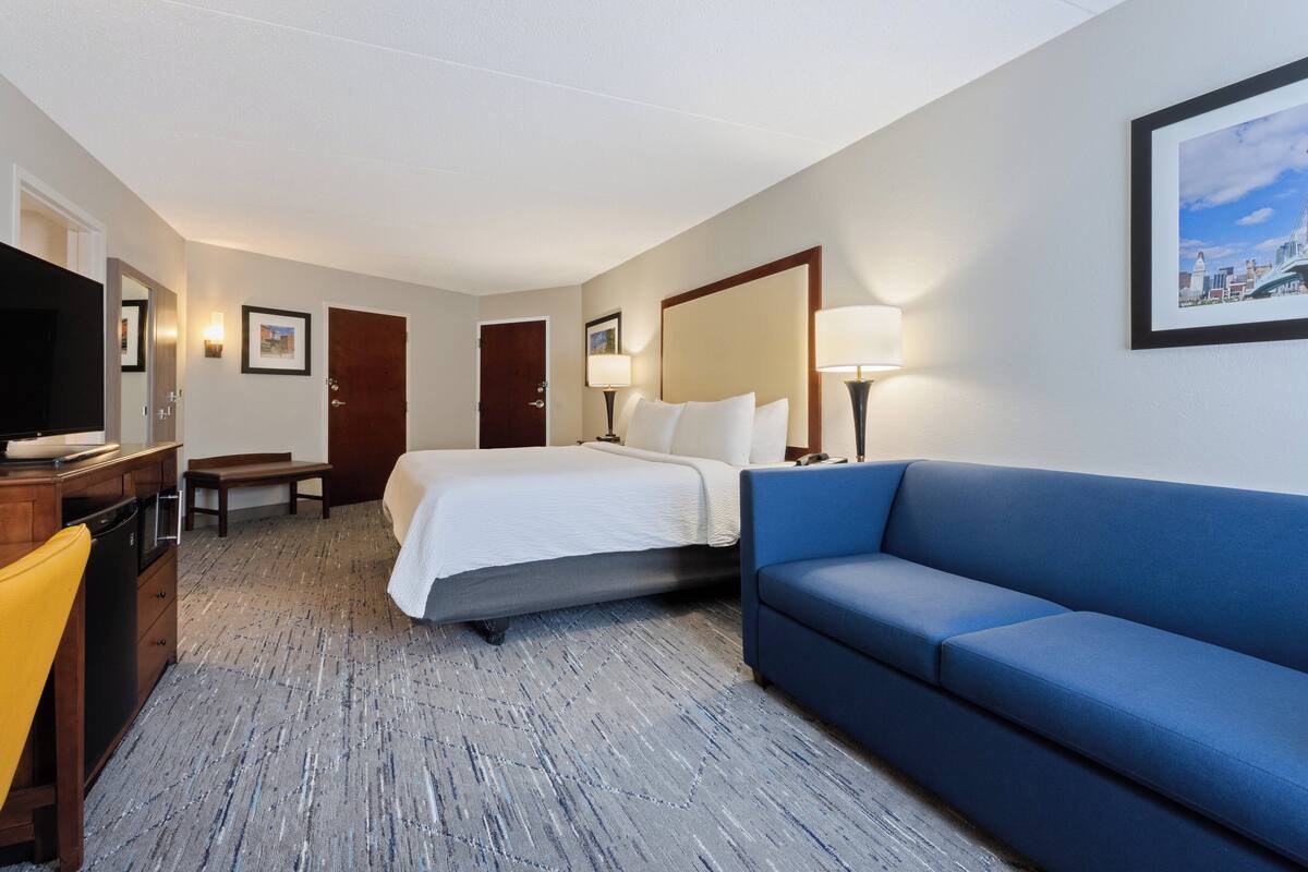 A hotel room with a bed, a blue sofa, a desk with a television, and a mirror.