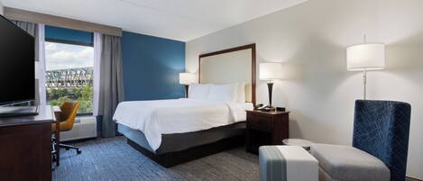 Habitación de hotel con cama, escritorio con televisor, silla, lámpara y vista a un puente.