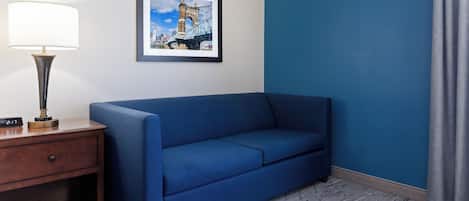 Una habitación con un sofá azul, una mesita de noche de madera con una lámpara, un cuadro con un puente y una pared azul.