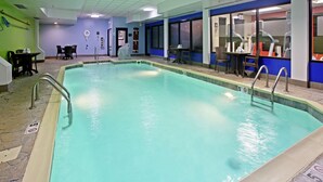 An indoor swimming pool with clear water, surrounded by a tiled floor and walls. There are chairs and tables around the pool area, and a door leading to another room.