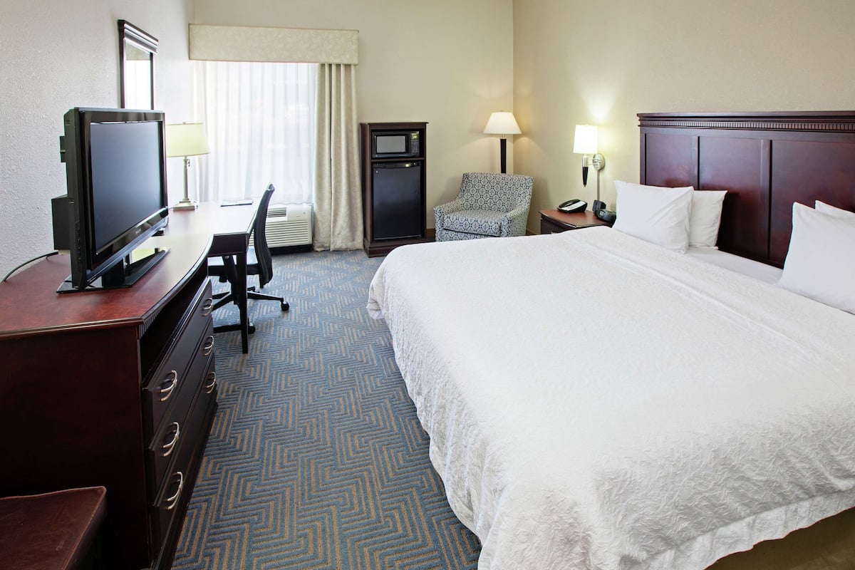 A hotel room with a large bed, a desk with a television, a chair, a desk lamp, and a nightstand with a phone.