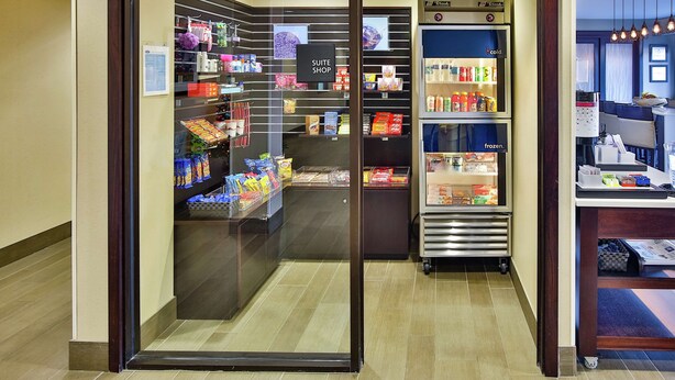 A hotel suite shop with shelves stocked with toiletries and snacks, a refrigerated section, and a visible exit sign.