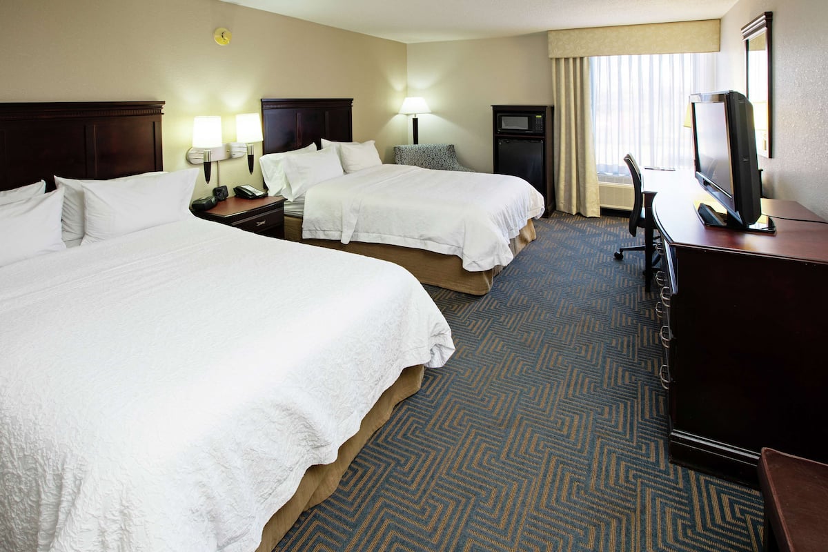 A hotel room with two beds, a desk with a television, a lamp, and a clock on the wall.