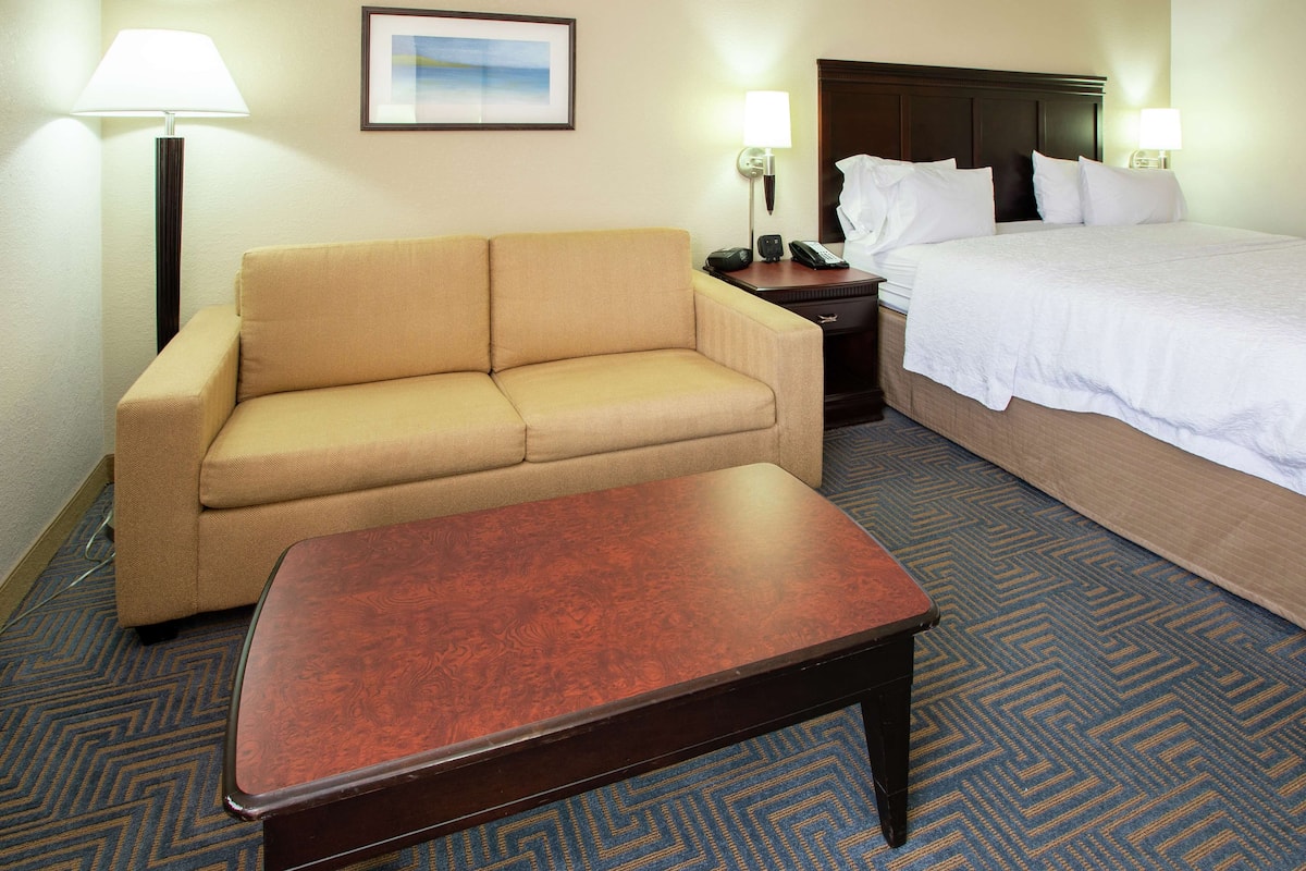 A hotel room with a beige sofa, a wooden table, a bed with white linens, a nightstand with a lamp, and a framed picture on the wall.