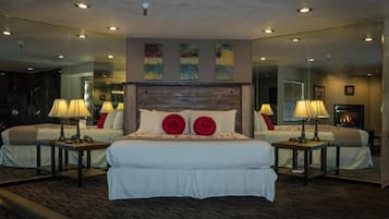 Una habitación de hotel con una cama grande, dos mesitas de noche con lámparas, una chimenea y paredes espejadas.