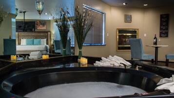 A hotel room with a large bathtub, a bed with blue bedding, a mirror, a window with blinds, a small table with a bottle and glass, and a chair.
