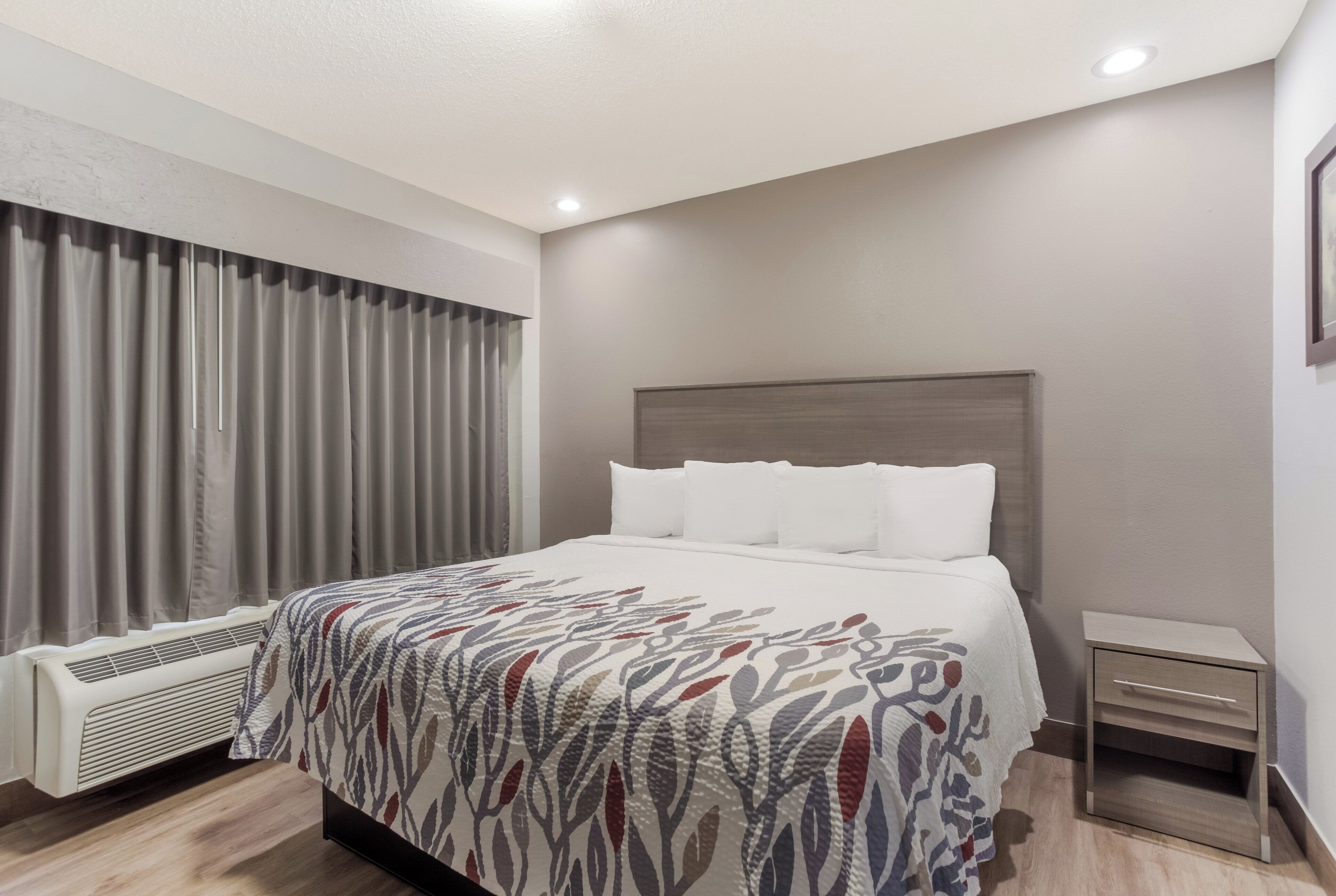Quarto de hotel com cama, mesa de cabeceira, aquecedor e janela com cortinas.
