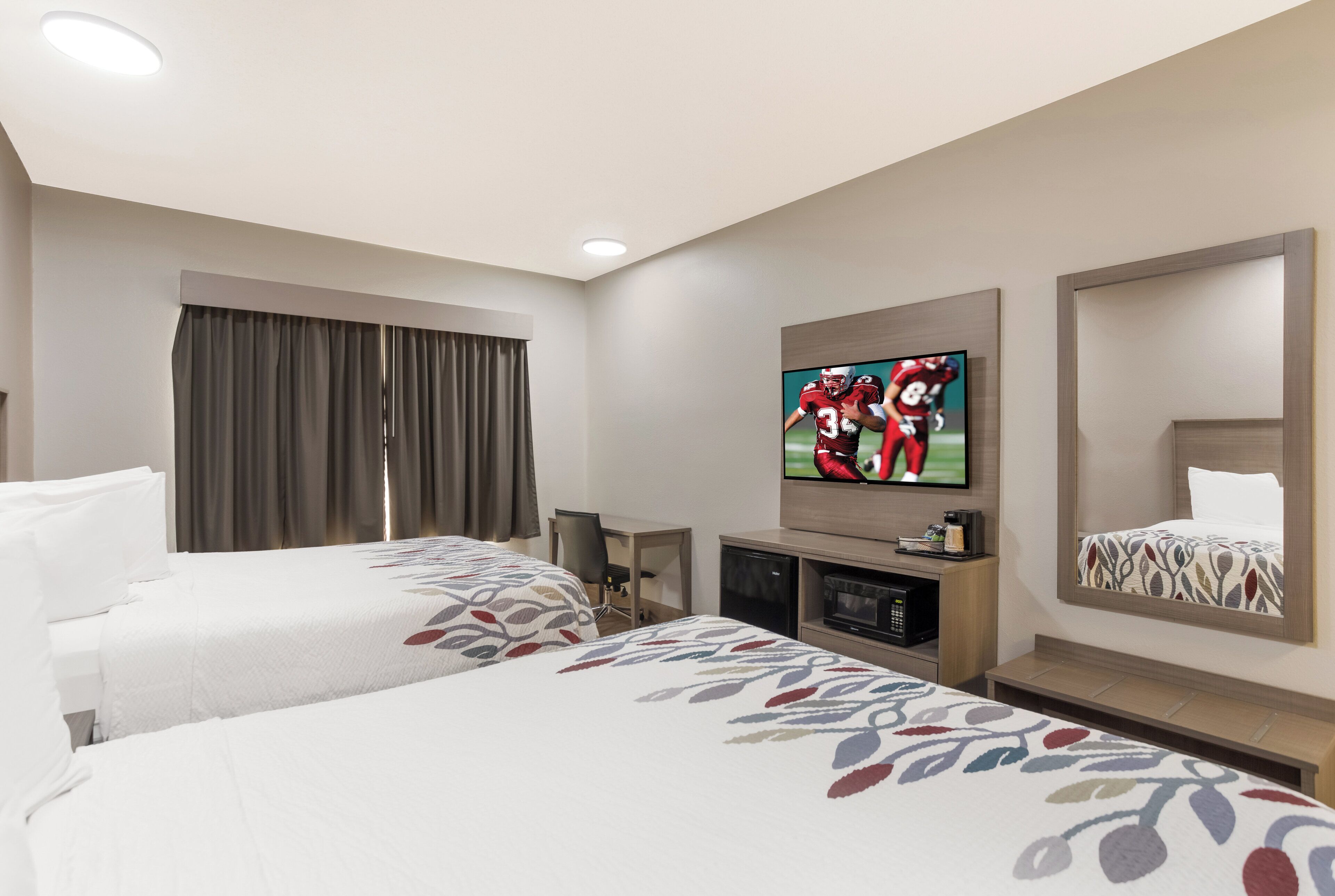 A hotel room with two beds, a television, a desk with a chair, a microwave, and a large mirror.