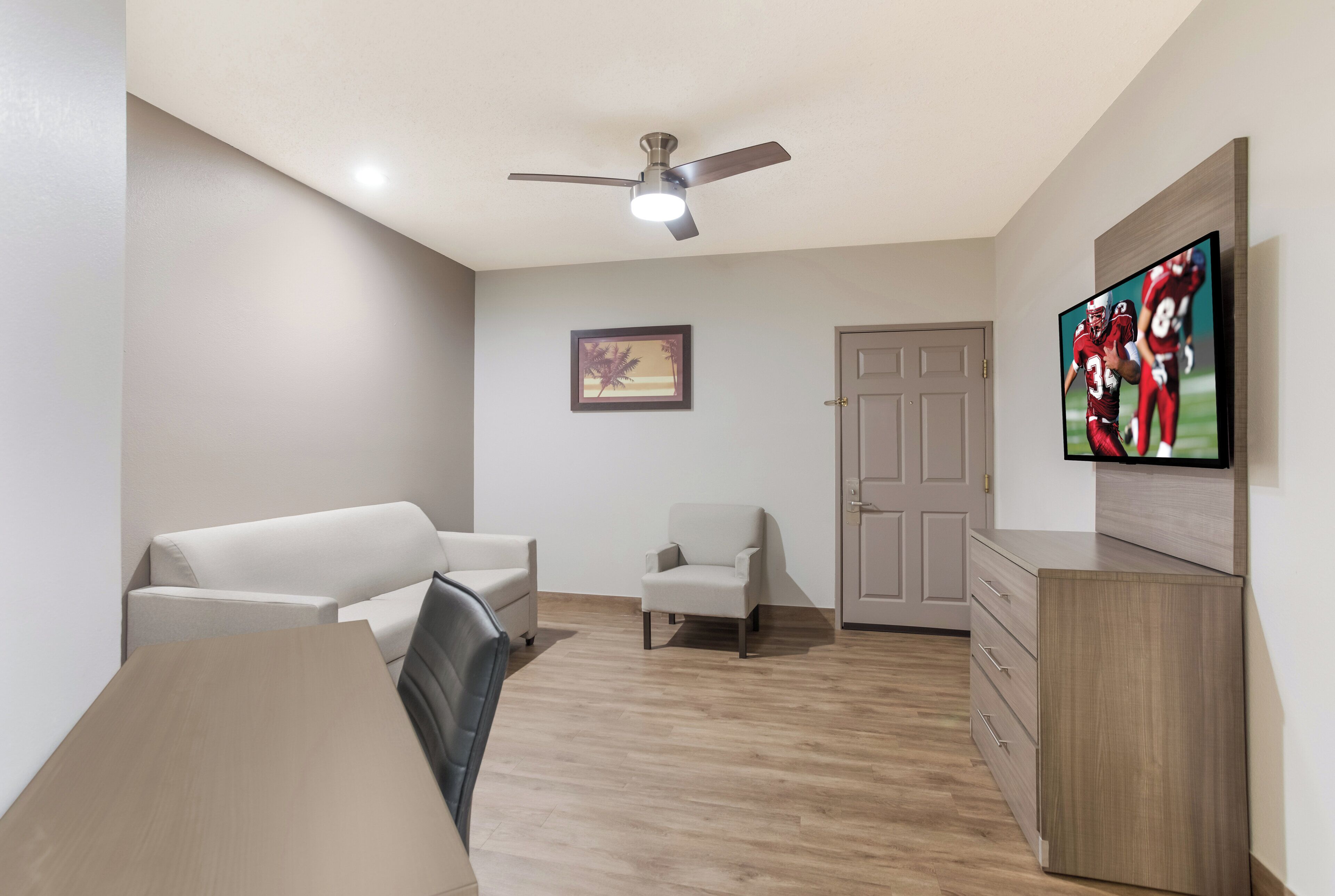 A room with a ceiling fan, a television mounted on the wall, a door, and a small framed picture.