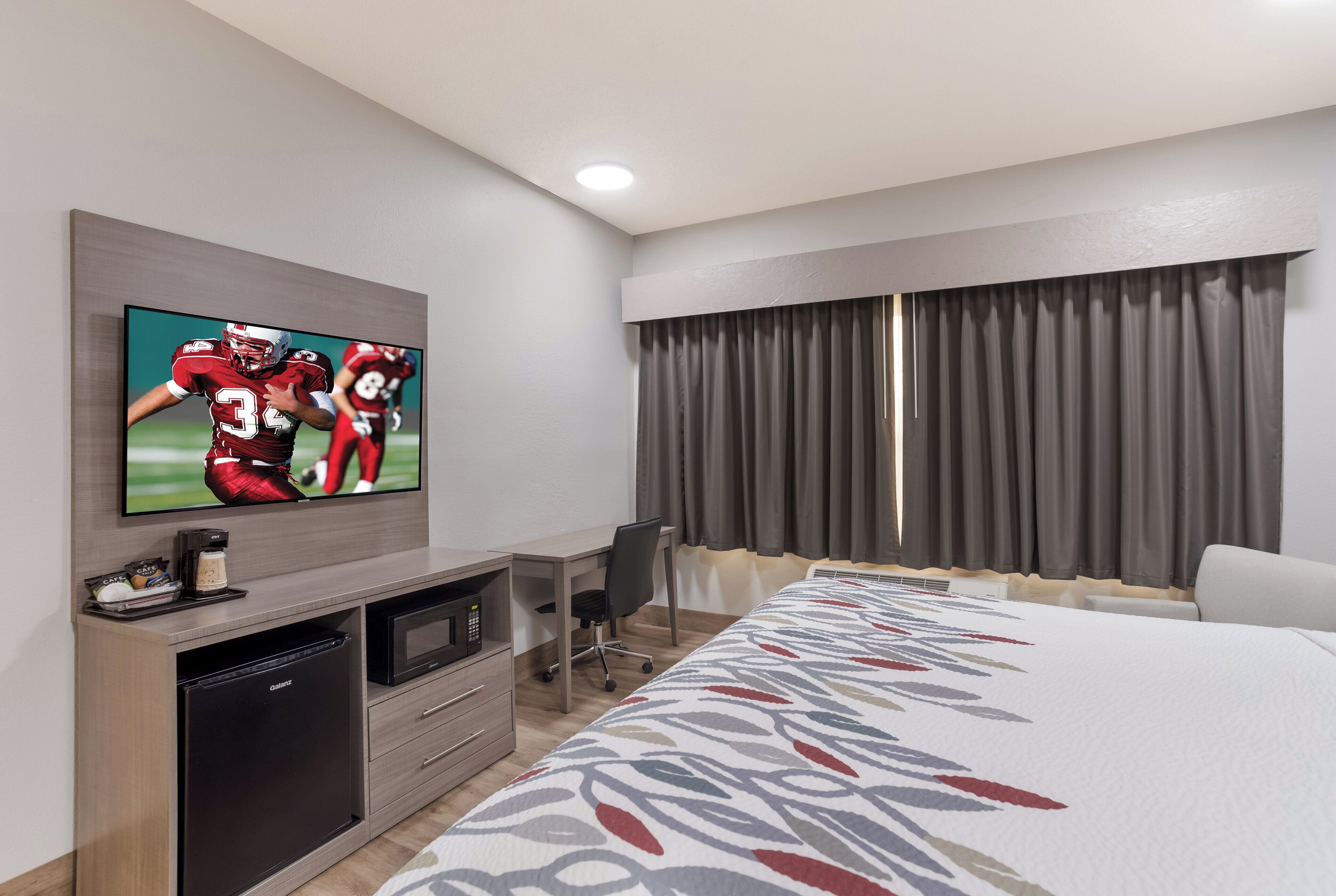 A hotel room with a flat-screen TV mounted on the wall, a desk with a microwave, a chair, a bed with a patterned bedspread, and a large window with curtains.
