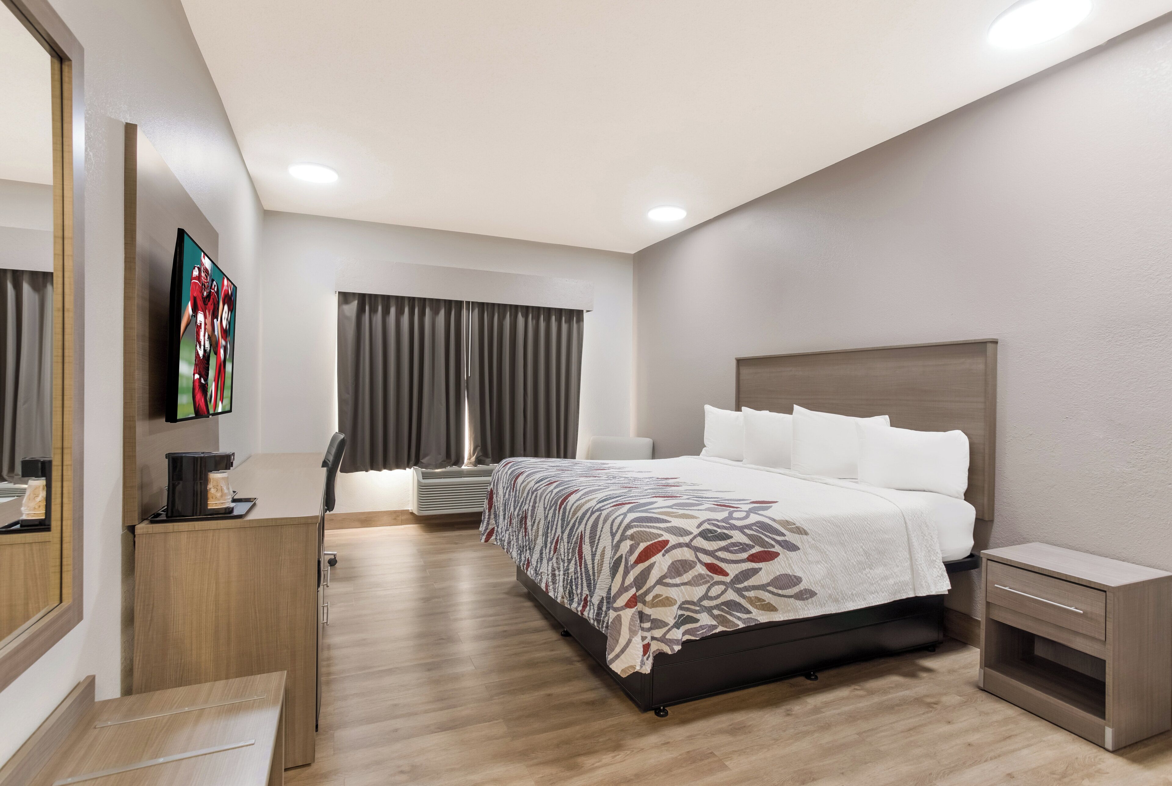 A modern hotel room with a large bed, a flat-screen TV, a desk with a coffee maker, and a window with curtains.