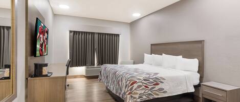 A modern hotel room with a large bed, a flat-screen TV, a desk with a coffee maker, and a window with curtains.