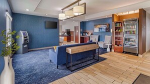 Un moderno hall de hotel con un sofá azul, un banco de madera, un refrigerador con bebidas y un quiosco de autoservicio.
