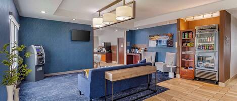 A modern hotel lobby with a blue sofa, a wooden bench, a refrigerator with beverages, and a self-service kiosk.