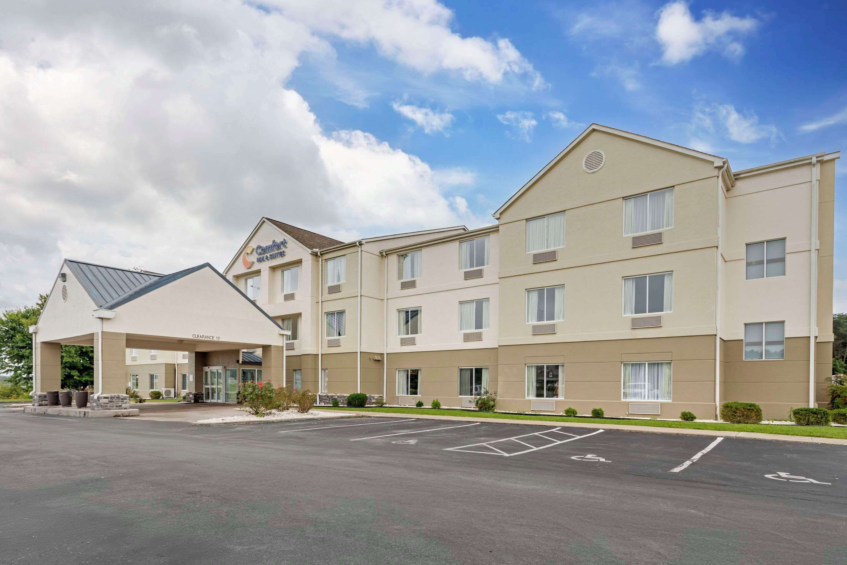 A multi-story hotel with a covered entrance, parking lot, and a clear sky.