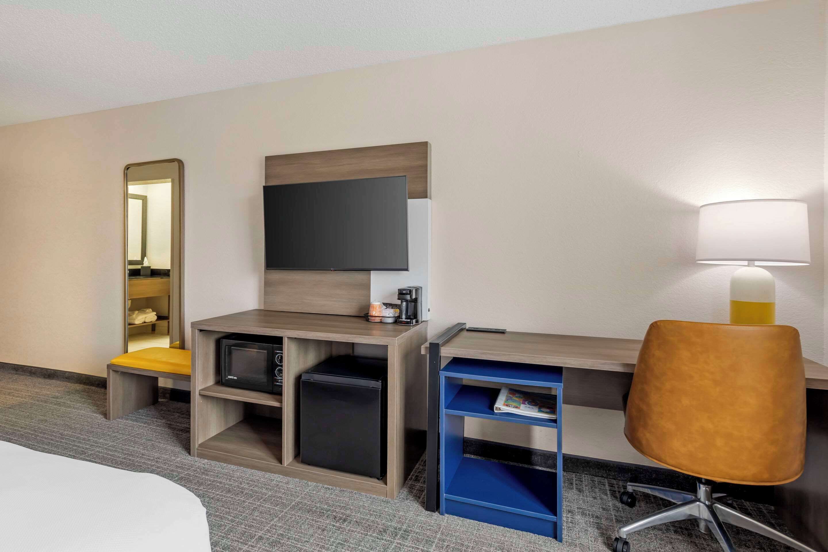 A hotel room with a bed, a desk with a computer, a chair, a TV, a lamp, and a small fridge.