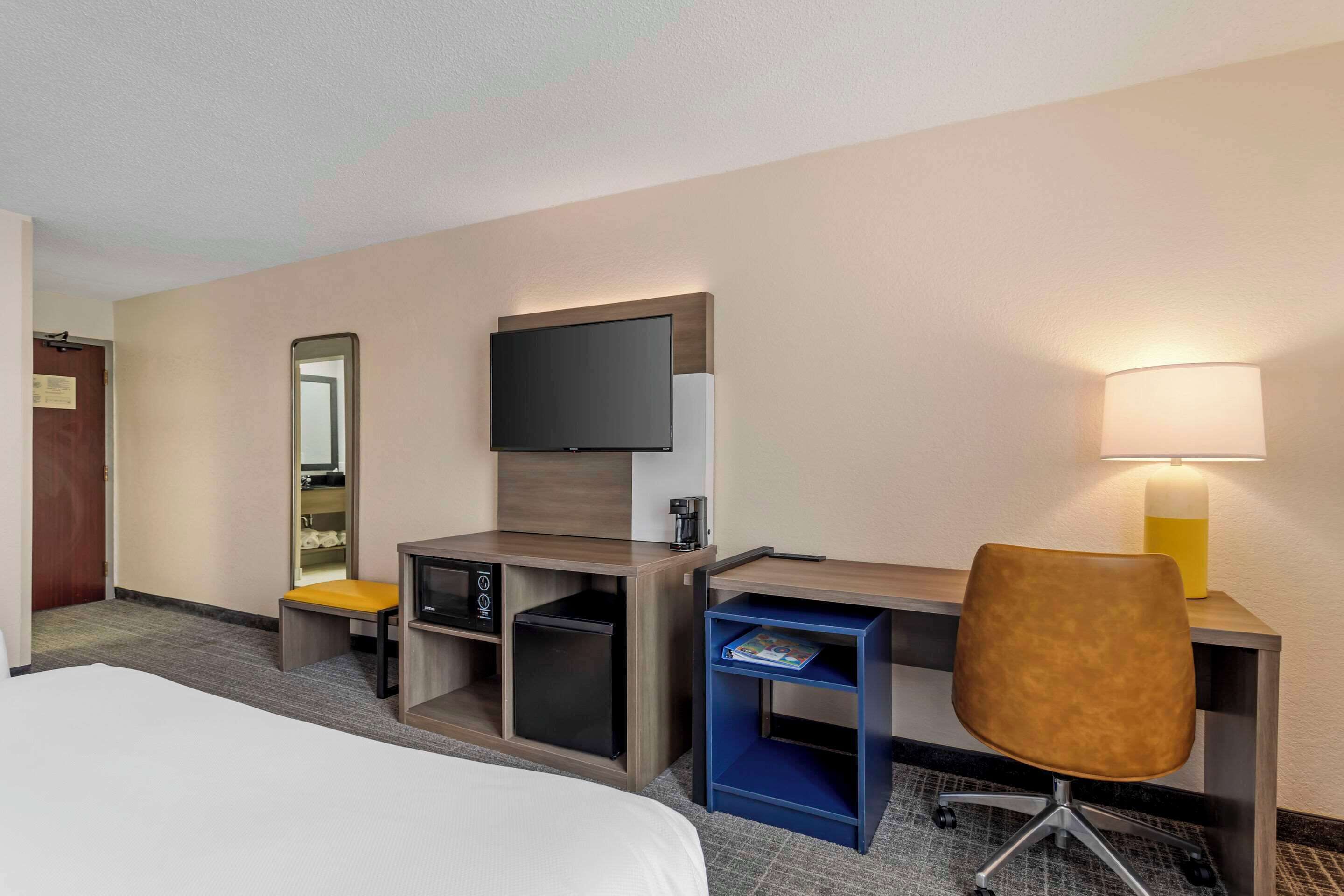 Habitación de hotel con una cama, un escritorio con ordenador, una silla, televisión, lámpara y estante pequeño.