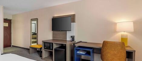 Habitación de hotel con una cama, un escritorio con ordenador, una silla, televisión, lámpara y estante pequeño.