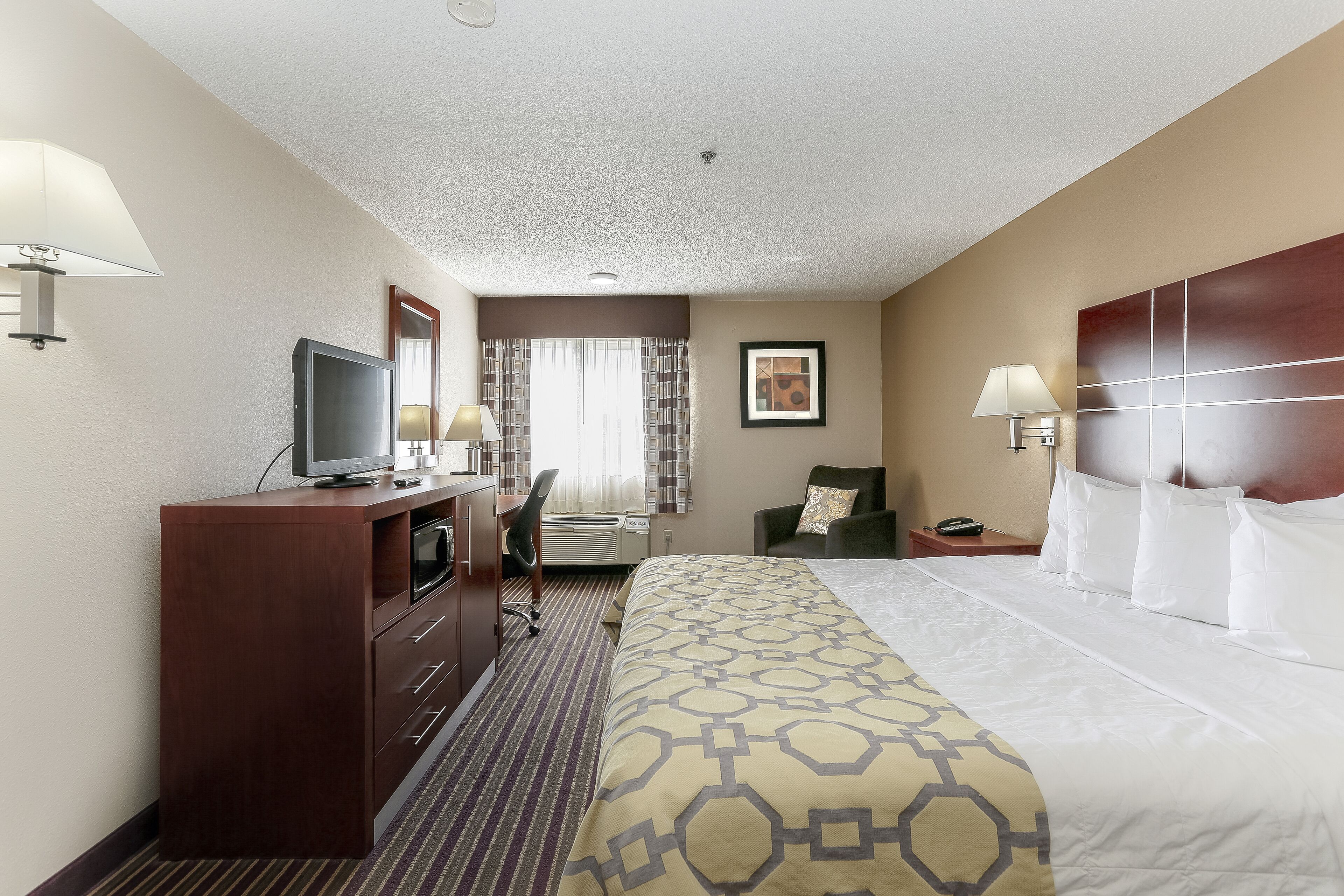 A hotel room with a bed, a desk with a TV, a chair, a nightstand, and a lamp.