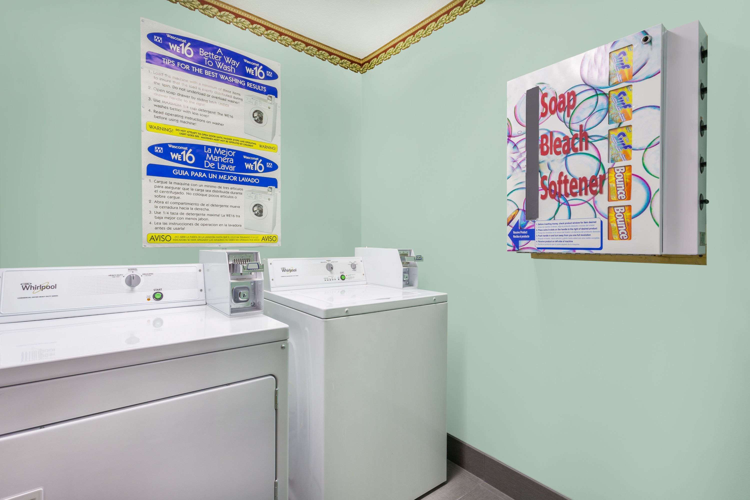 A laundry area with two Whirlpool washers and dryers, a wall-mounted instruction poster, and a soap advertisement.