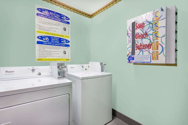 A laundry area with two Whirlpool washers and dryers, a wall-mounted instruction poster, and a soap advertisement.