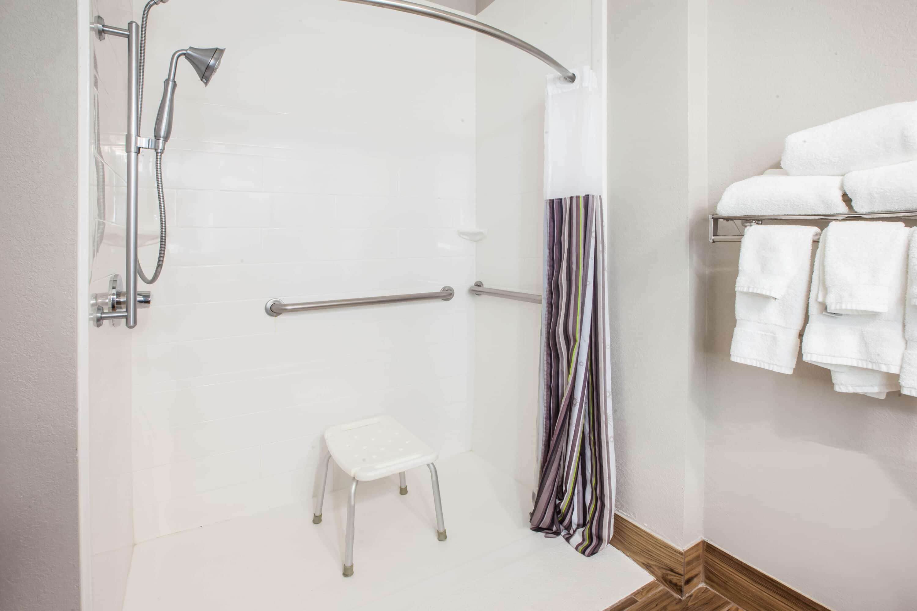 Une douche équipée d’un pommeau de douche à main, d’un petit tabouret blanc et d’un porte-serviettes avec des serviettes pliées.