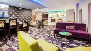 Un moderno vestíbulo de hotel con alfombra estampada, un sofá morado, sillones amarillos y una zona de comedor con mantel de estampado en zigzag.