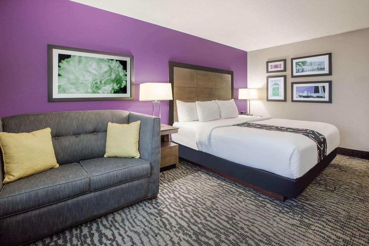 Habitación de hotel con sofá gris, cama grande con ropa blanca, cabecero, mesita de noche, lámpara y un cuadro en la pared.