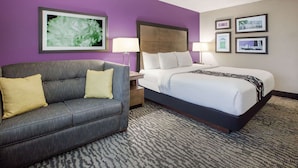Habitación de hotel con sofá gris, cama grande con ropa blanca, cabecero, mesita de noche, lámpara y un cuadro en la pared.