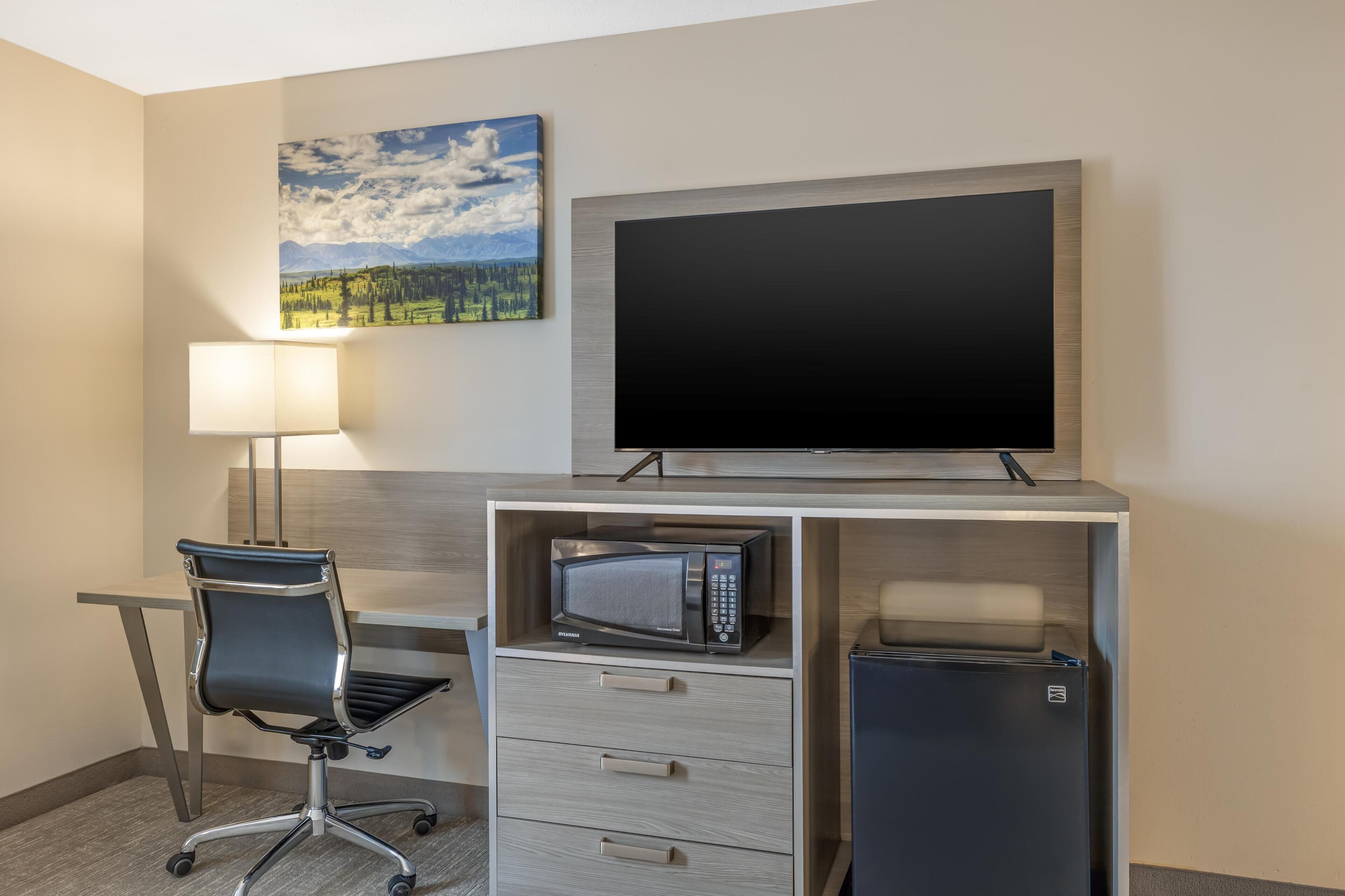 Habitación de hotel con escritorio, silla, televisión, microondas, refrigerador y un cuadro en la pared.